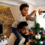 A young boy sits on his father's shoulders while he places a star on top of a Christmas tree covered in ornaments
