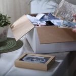 A white box sits on a table with a photo frame and photos next to it as an older person sorts through the box of old letters and belongings.