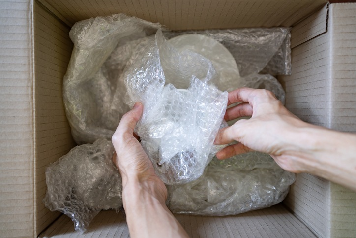 A pair of hands are wrapping fragile pieces of dinnerware in bubble wrap before placing them in a cardboard box.