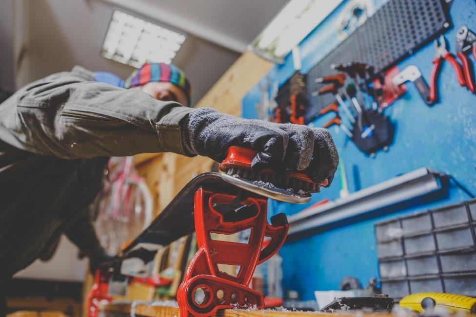 A man wearing a long sleeve shirt and gloves cleans the wax off a dark colored ski. The ski rests on a red metal support and the brush being used is red as well.