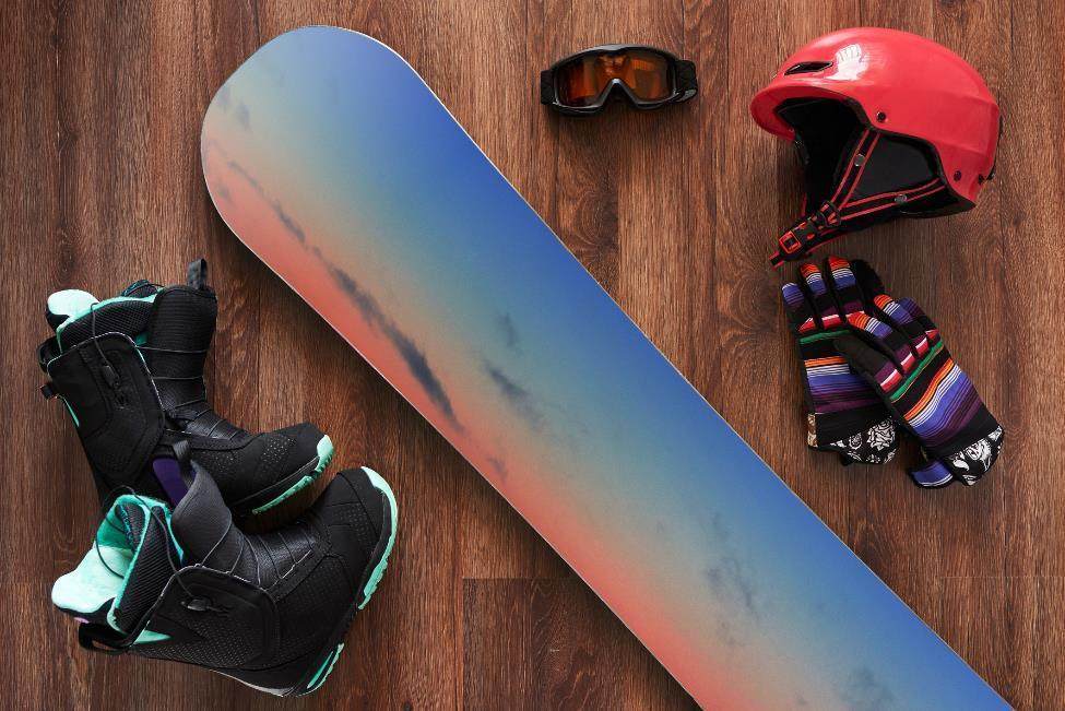 A blue, yellow, and pink snowboard rests on a wooden floor. It is surrounded by a red helmet, gloves, and black ski boots.