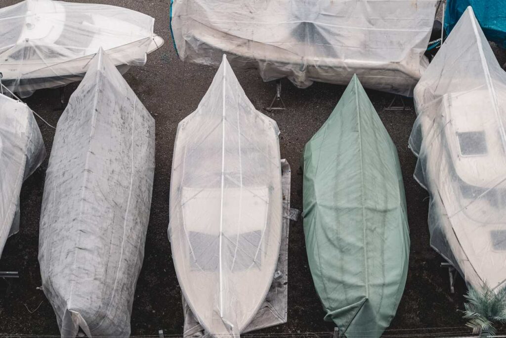 A line of boats are covered and sit in a row in an outdoor lot.