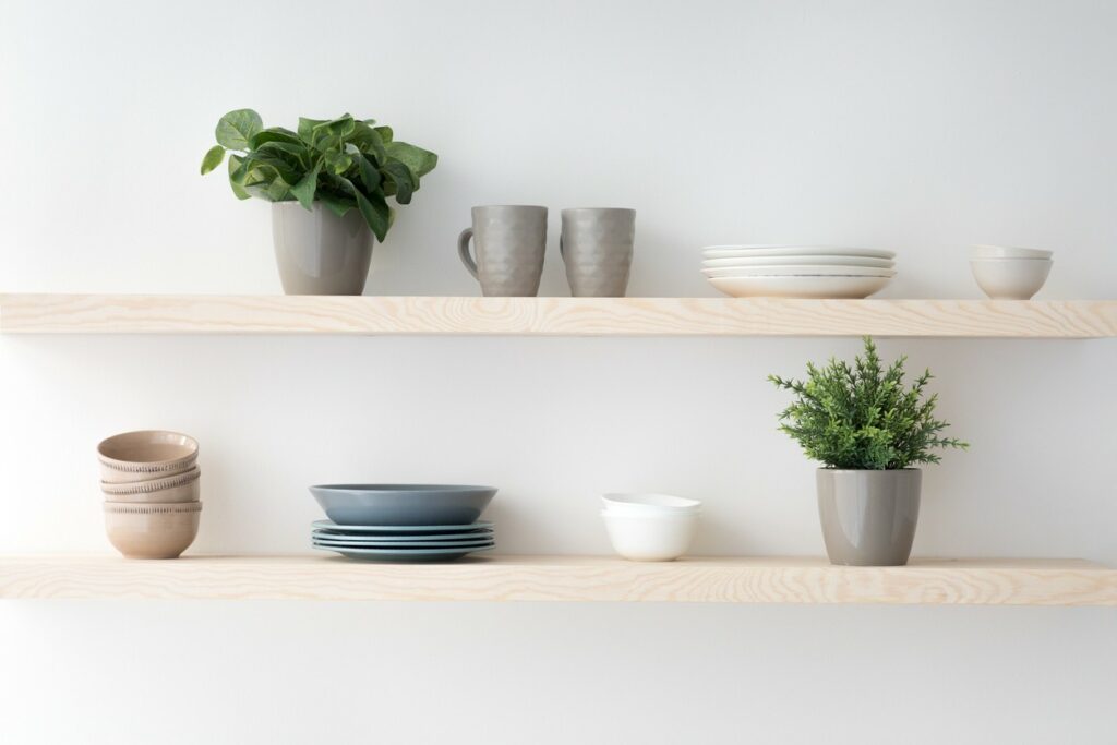 Two wooden shelves sit on a wall and hold plates, cups, bowls and two plants.