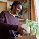 A young man wearing a purple sweater unpacks a plant from a box in a dorm room