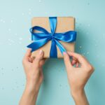 A person holds a package wrapped in brown paper and a blue bow above a blue background with glitter.