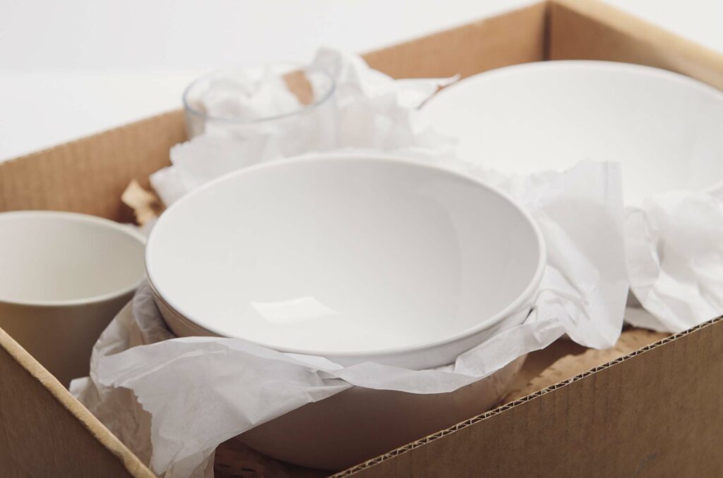 A bowl and packing materials on top of a variety of dishes in a box.