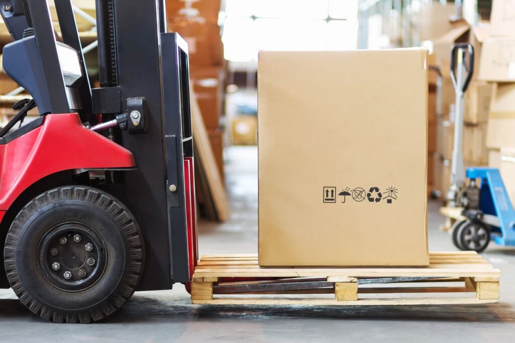 Part of a forklift in the form of a wheel and a pitchfork holding a large box.