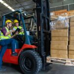 Two workers working together to carry boxes on a forklift in a warehouse.