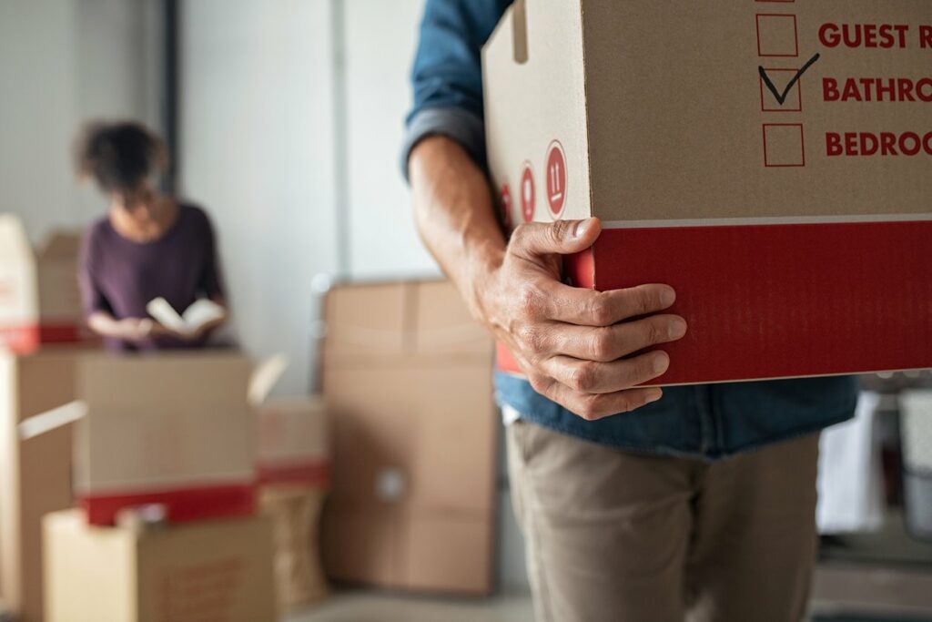 A man holding a cardboard box with woman packing items in the background. 