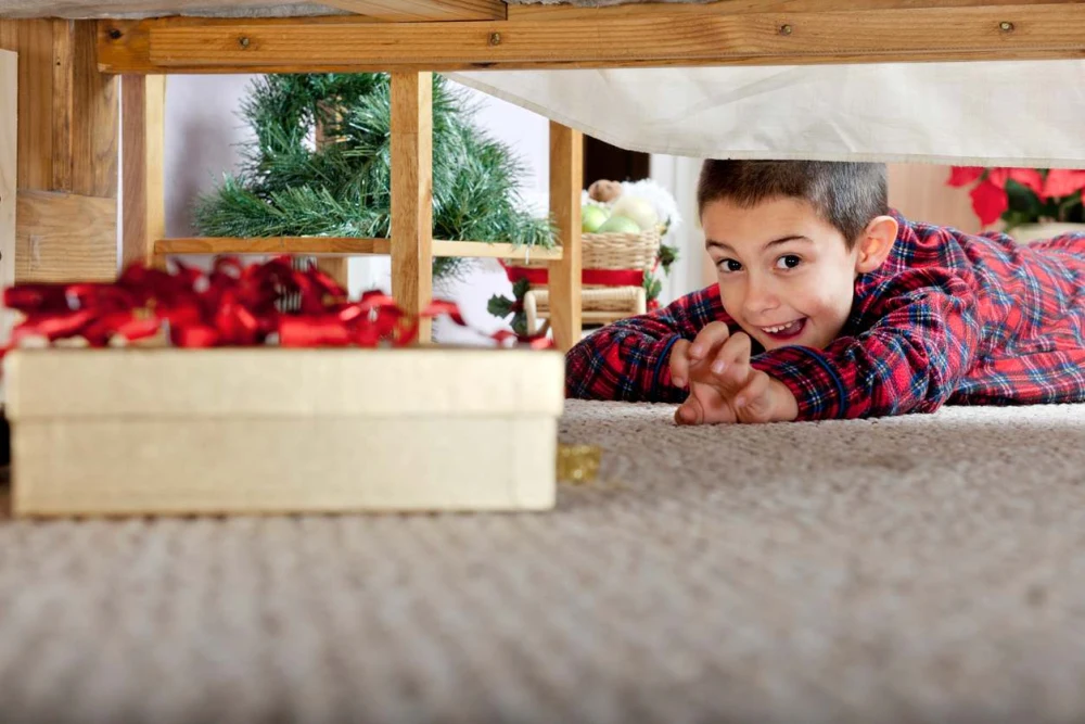 A young child reaches for a gift box underneath a bed. 