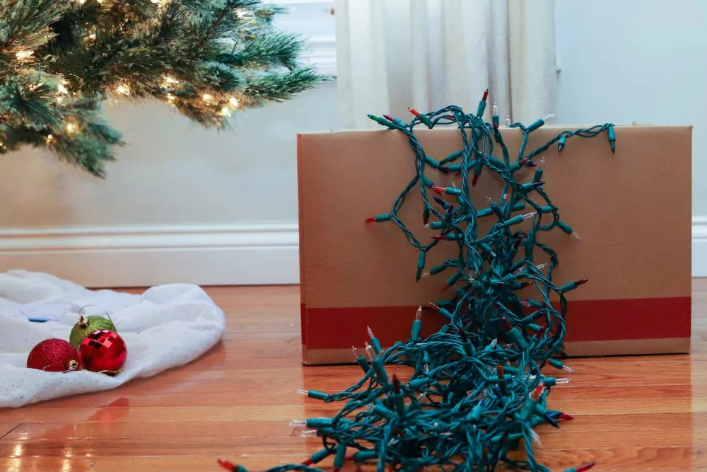 String lights are draped over a cardboard box next to a Christmas tree. 