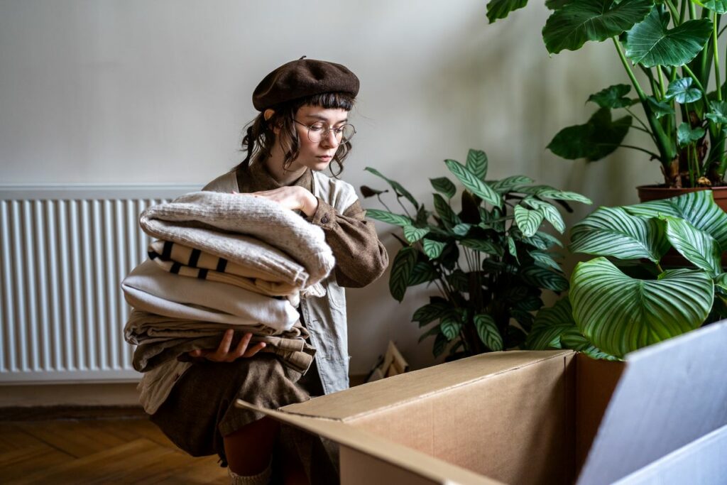 Woman placing folded piles of clothing in cardboard box for decluttering.