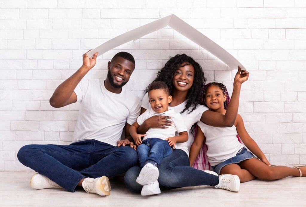 A family sitting under a folded board that they’re holding to resemble their new home. 