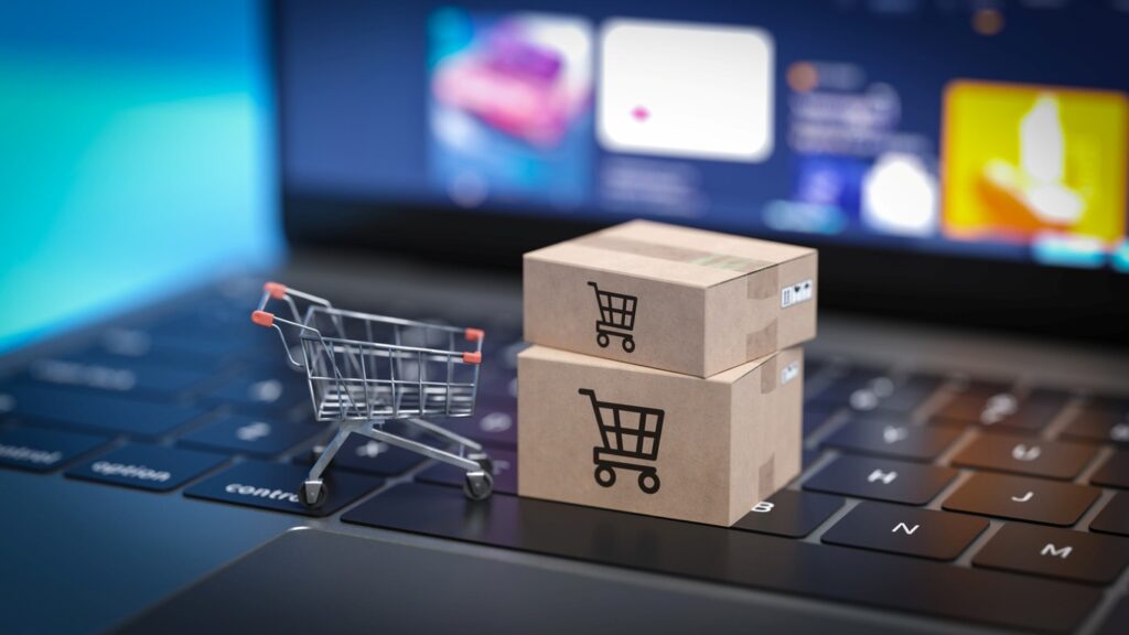 A mini shopping cart and miniature cardboard boxes sit on a laptop keyboard to resemble e-commerce. 