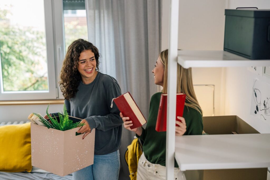 Two college students pack up their dorm room together, smiling as they box books and plants for storage.